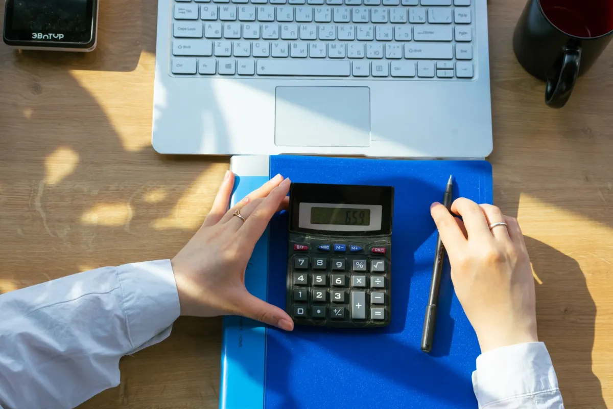 Hands using a calculator on a blue folder in front of a laptop
